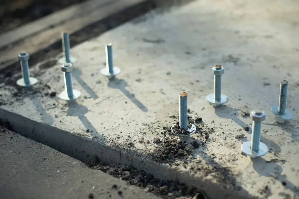 Kilka metalowych kotew z podkładkami i nakrętkami osadzonych w betonie.